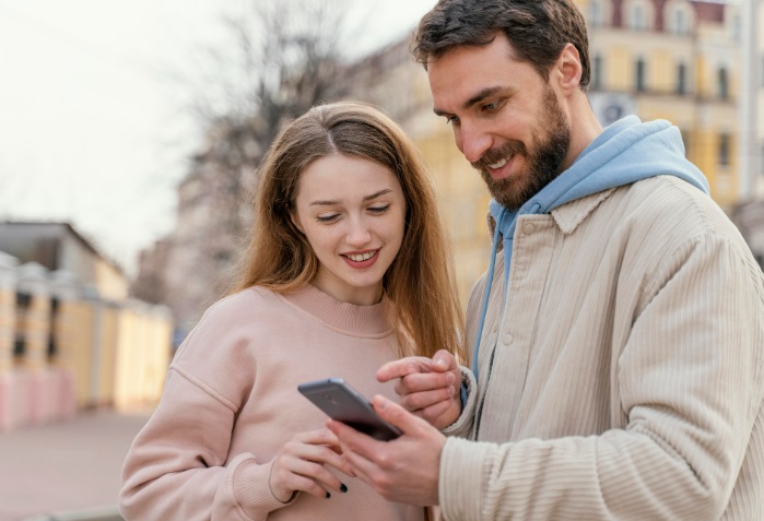 Deux personnes souriantes regardent un téléphone à l’extérieur